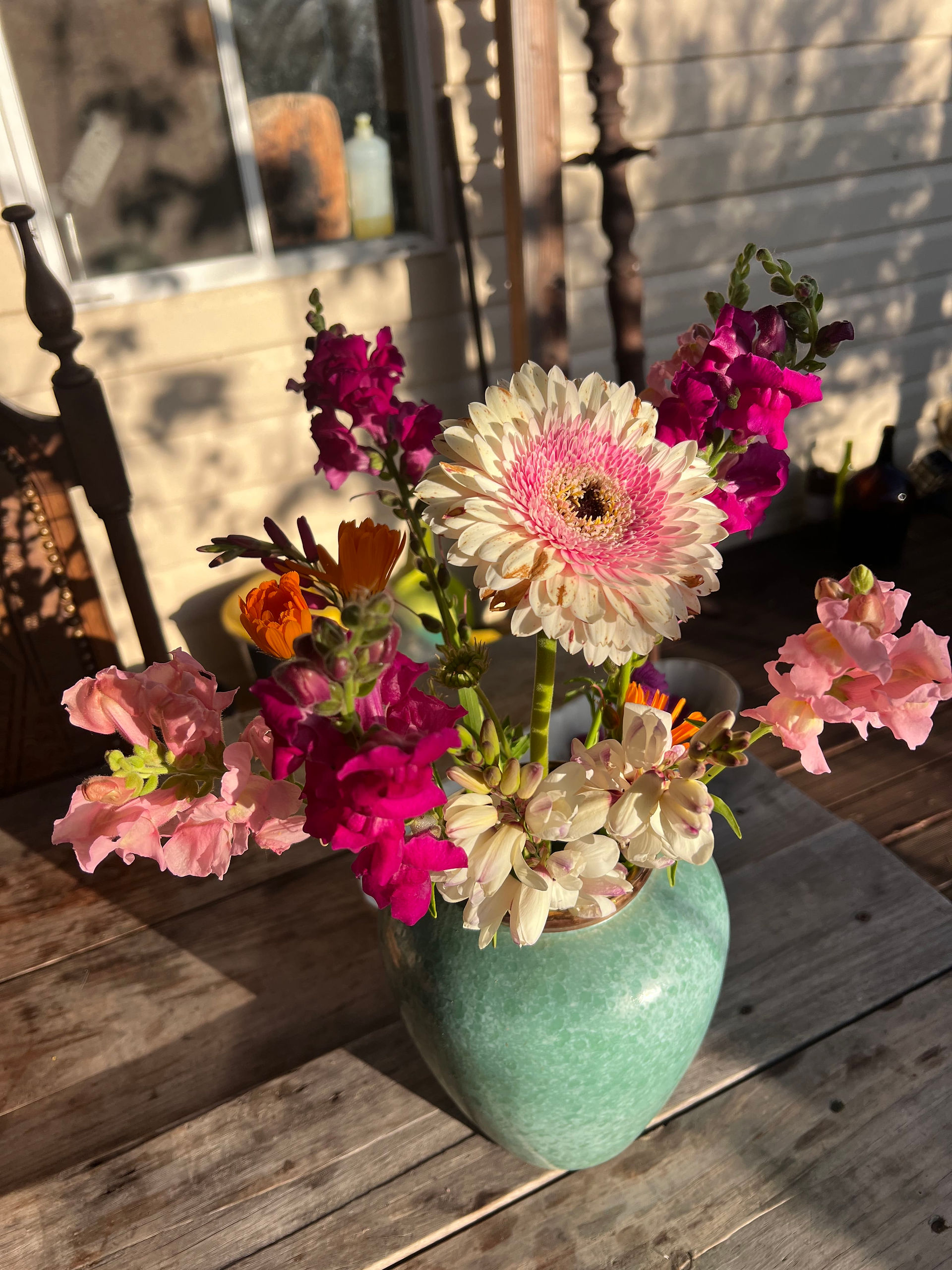 A pink and white Gerbera daisy arranged with snapdragons and calendula in a green ceramic vase on a wooden table.