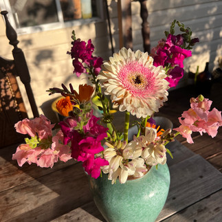 A pink and white Gerbera daisy arranged with snapdragons and calendula in a green ceramic vase on a wooden table.