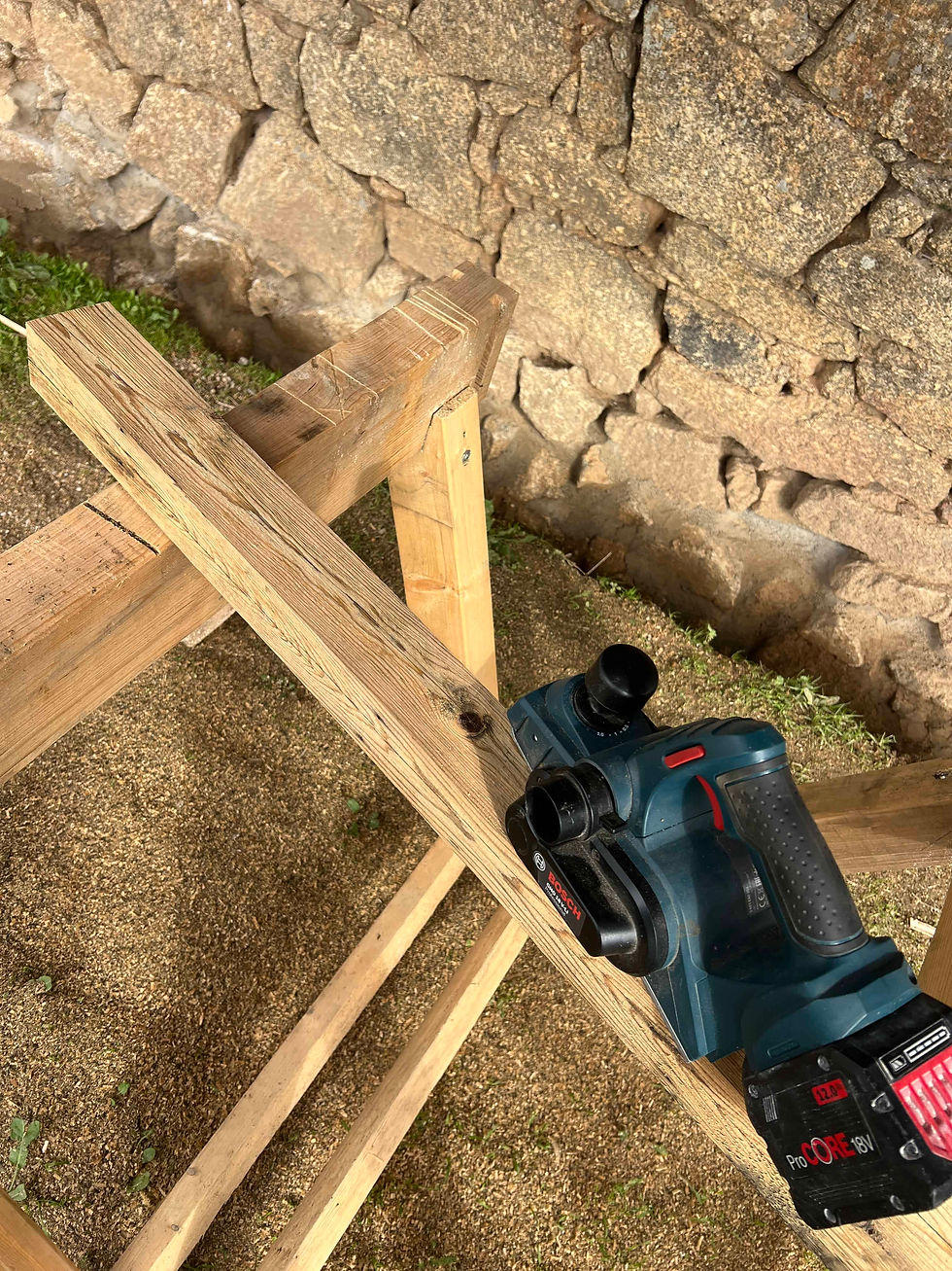 Handheld Bosch GHO 18V-LI planer resting on a wooden beam supported by a simple sawhorse near a stone wall.