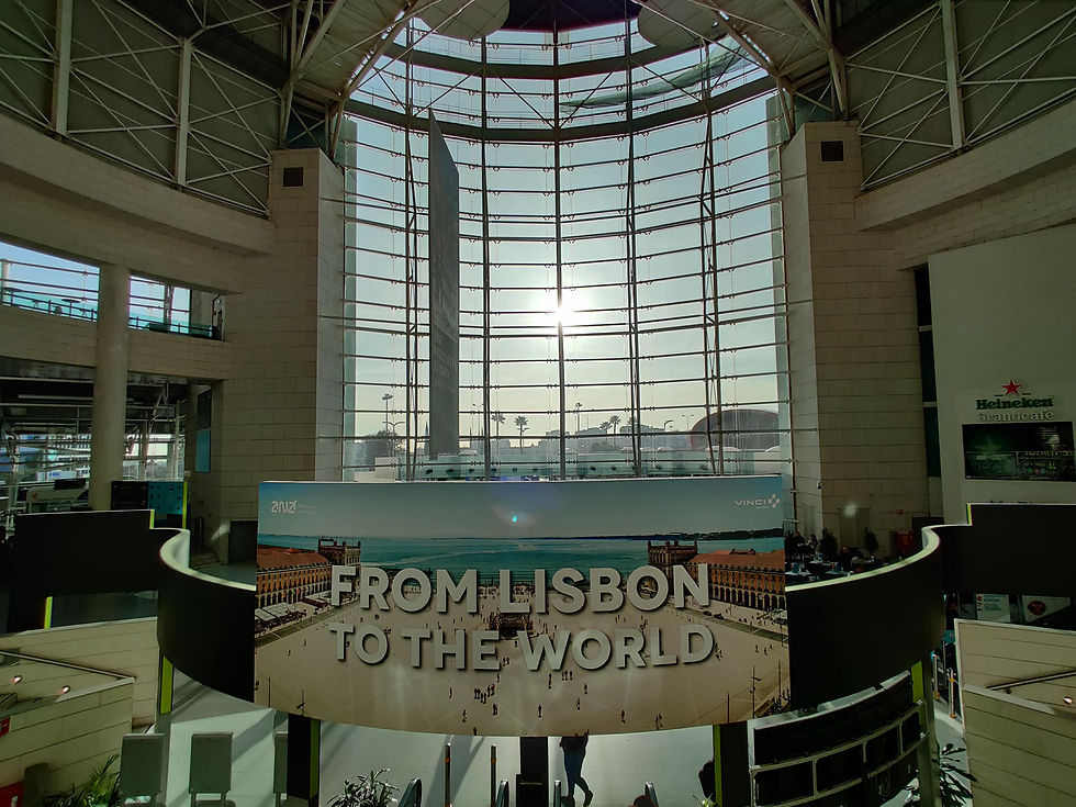 Large airport atrium with tall glass windows and sunlight shining through above a sign reading “From Lisbon to the World.”