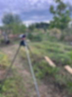 Tripod-mounted Gardena Pulse sprinkler stands on a garden path beneath a cloudy sky, overlooking grassy beds, young trees, and scattered shrubs.
