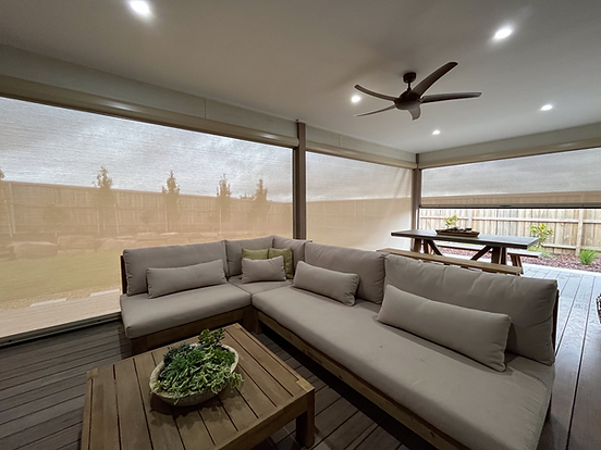 An outdoor patio area with beige ziptrak blinds, a sofa with cushions, a wooden coffee table with a plant, a picnic-style dining table, and ceiling fans.
