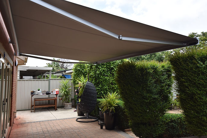A modern retractable awning extends over a patio, creating a shaded outdoor space with lush green plants and a comfortable hanging chair.
