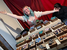 Stand de dés artisanaux de La Looterie en convention, avec une exposante accueillante devant une grande variété de sets de dés présentés dans des boîtes en bois