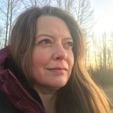 Shannon L. Post wearing a down cranberry jacket gazes thoughtfully into the distance, with bare trees and a setting sun in the background.