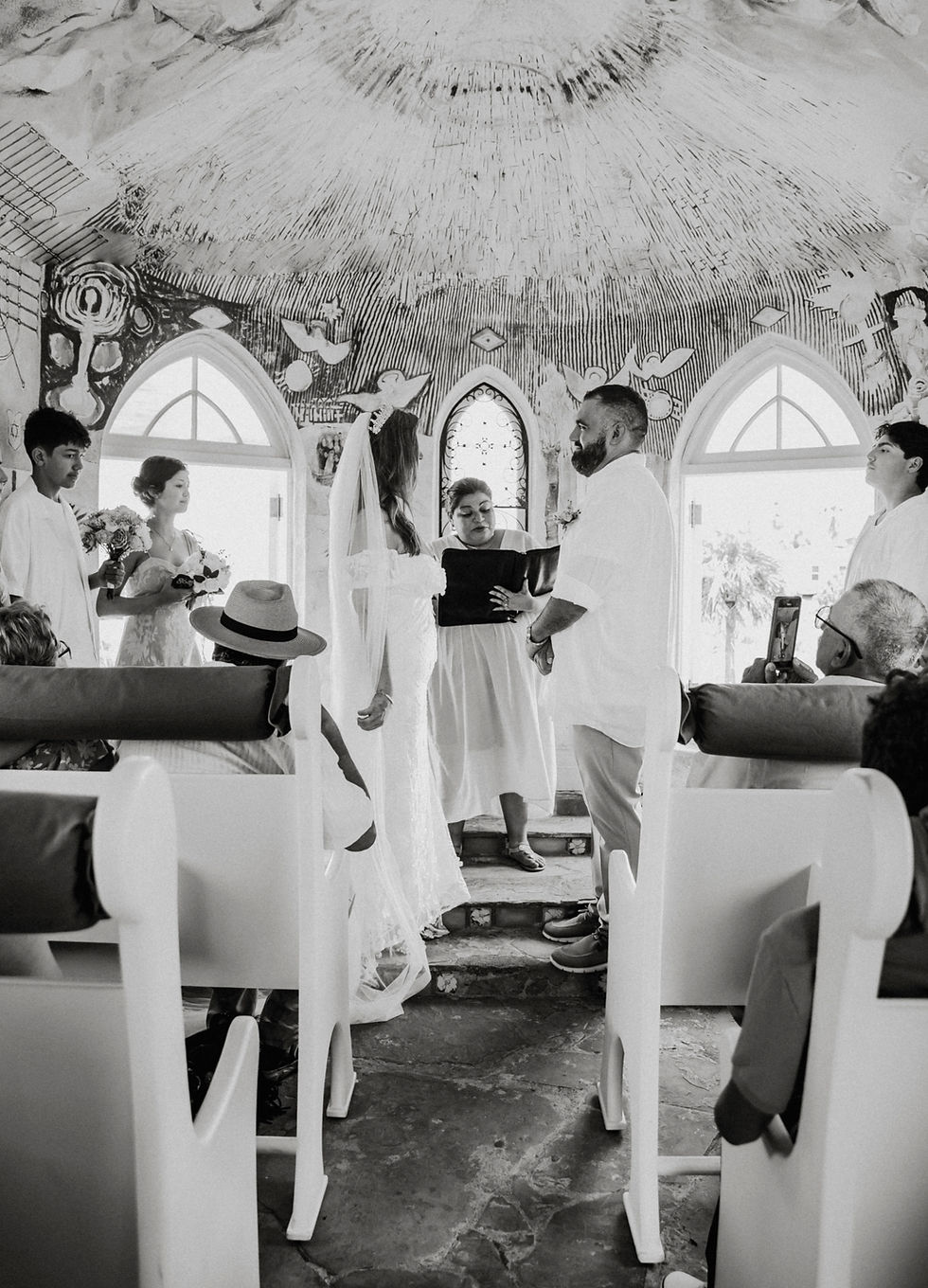 Bride and groom stand before officiant in ornate chapel, exchanging vows. Guests observe. Black and white image with artistic ceiling.