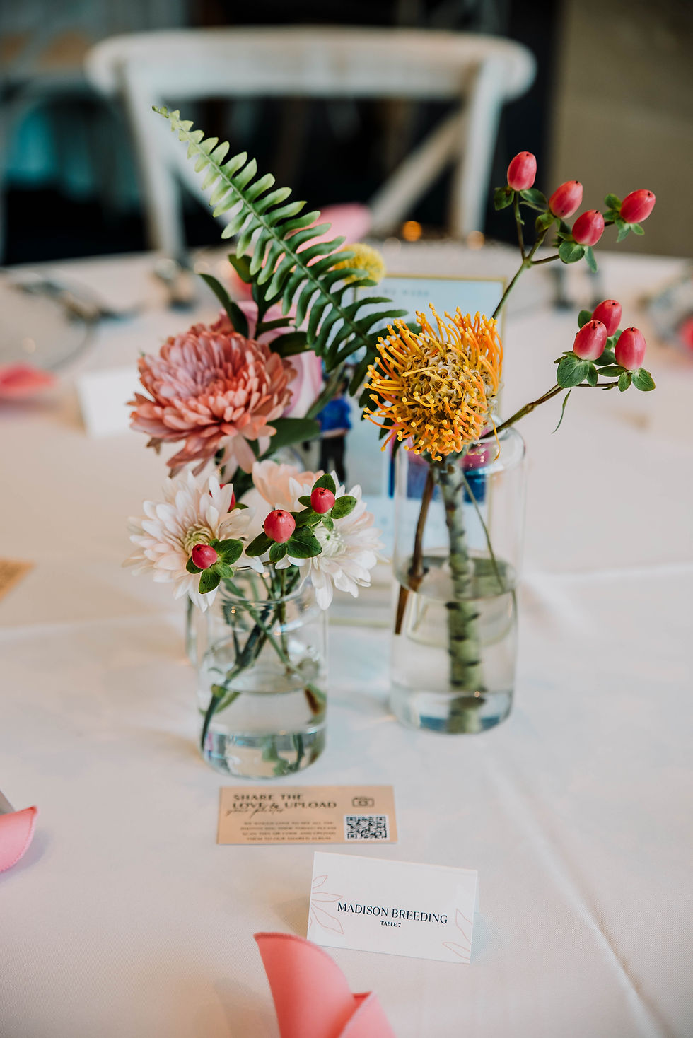 Colorful flowers in glass vases with a "Madison Breeding, Table 7" card on a white tablecloth, creating an elegant table setting.