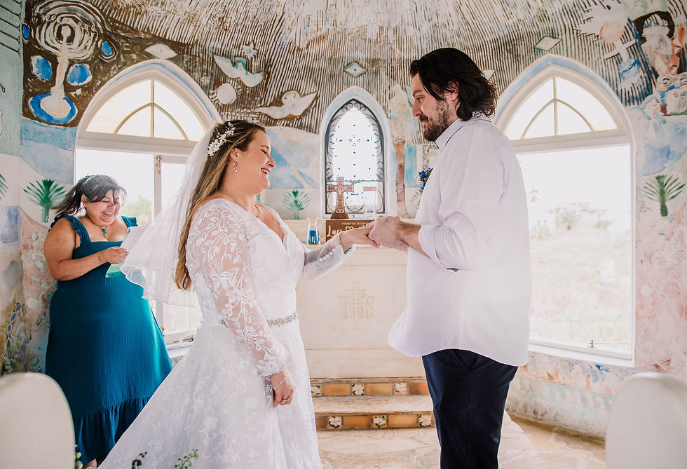 Bride and groom exchange rings in a colorful, artistic chapel. A woman in a blue dress smiles in the background.