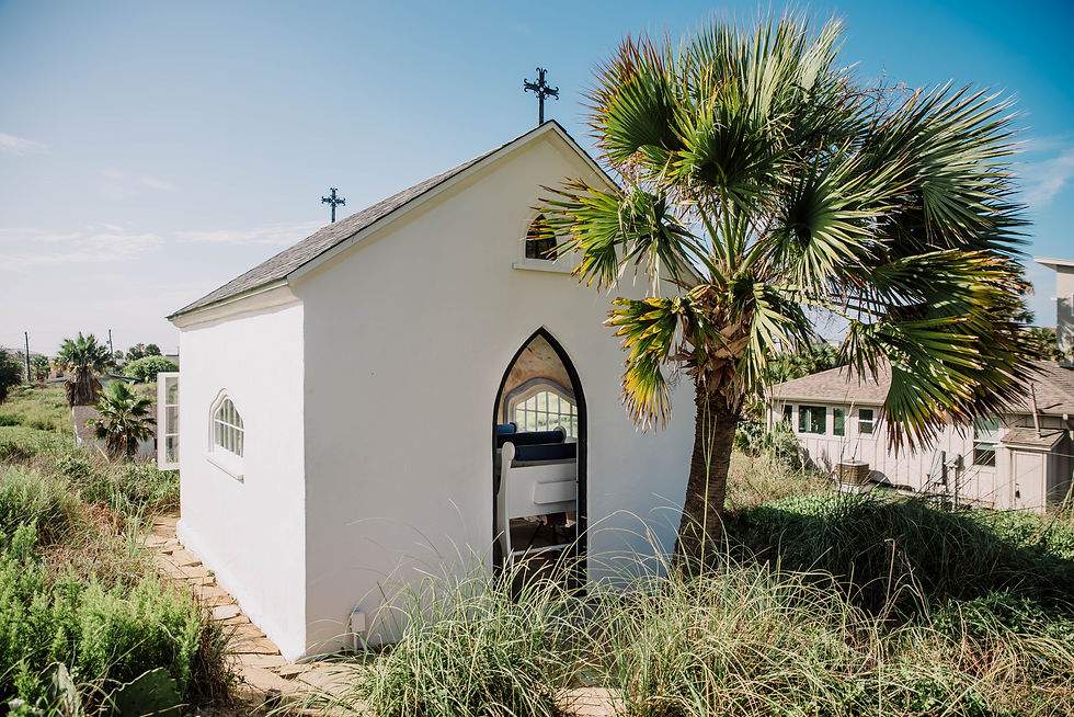 Small white chapel with arched window and cross, next to a palm tree under a clear blue sky. Grassy foreground, peaceful setting.