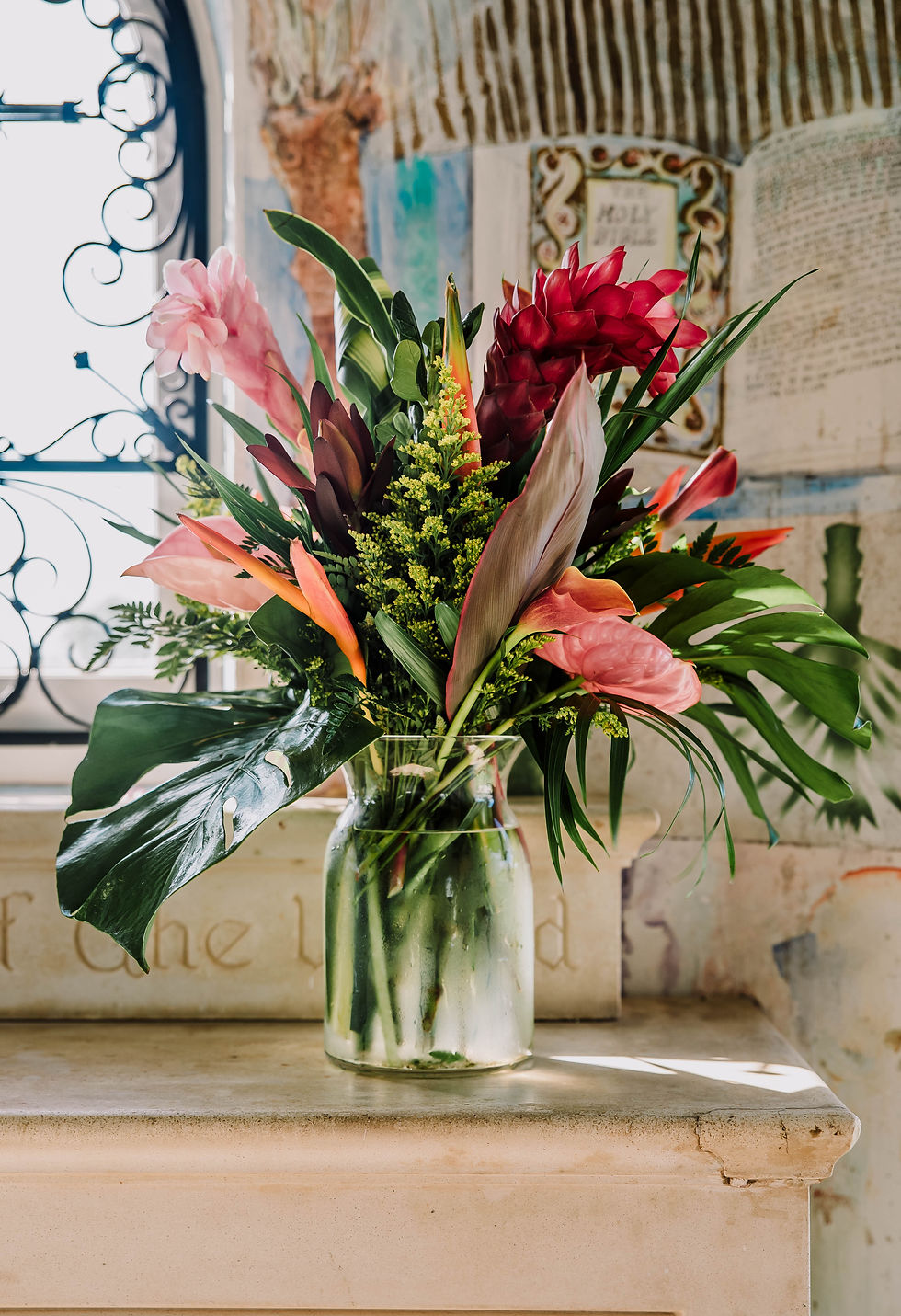 A vibrant bouquet with red, pink, and orange flowers in a glass vase sits on a sunlit table against a decorative, patterned wall.