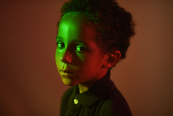 a boy with a serious look is bathed in green light with a reddish background.