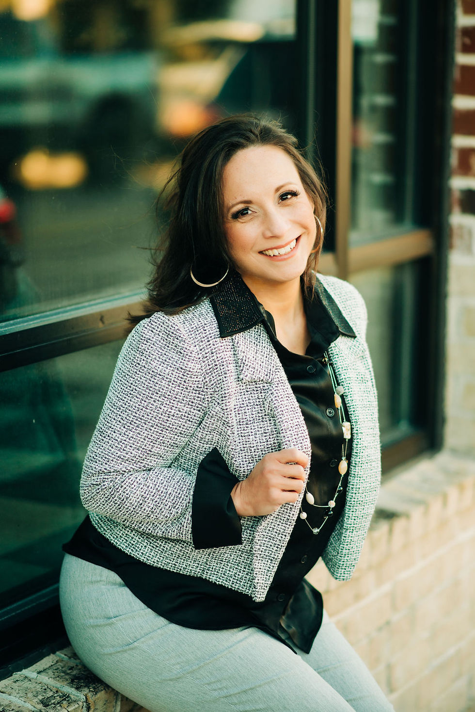 Portrait of a smiling woman sitting by a window, wearing a light textured blazer, black blouse, and light pants; warm tones, shallow depth of field, portrait orientation.