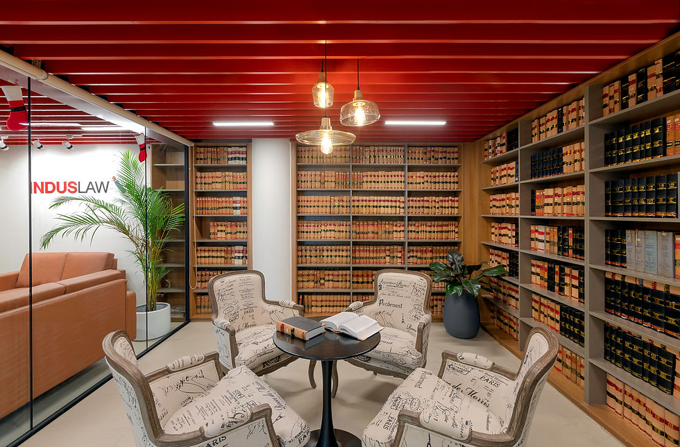 Library room with red ceiling, shelves of books, four patterned chairs, and a round table with open books. "INDUSLAW" text on glass wall.