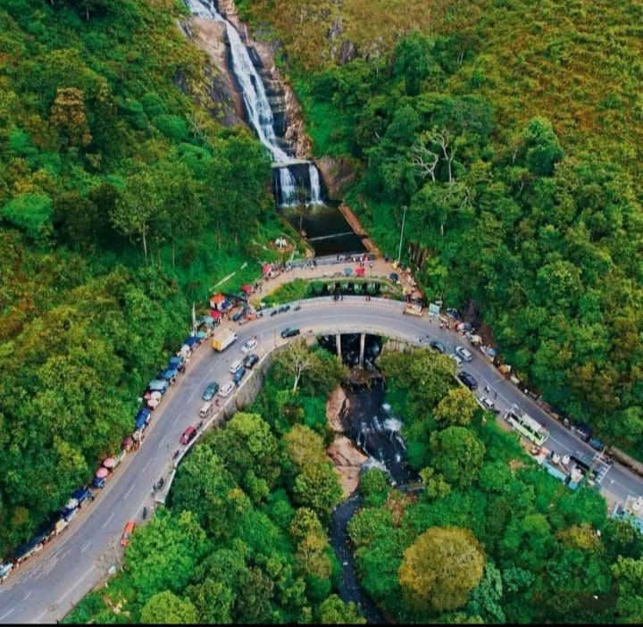 Aerial view of a lush green valley in Kodaikanal with a winding road curve, bridge, and tall waterfall cascading down the rocks