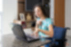 This image shows a young woman sitting in a wheelchair at a table using a laptop computer. She is using the computer with just her left index finger. She is wearing a light blue and white floral shirt.
