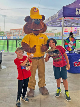 Two boys and Sam The Bear pose with baseballs in front of a tent.