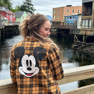 Ashlie wearing a mustard yellow plaid flannel with a large Mickey Mouse face on the back, standing on a wooden bridge in Ketchikan, Alaska — a cozy and playful outfit perfect for a Disney Alaska Cruise port day.