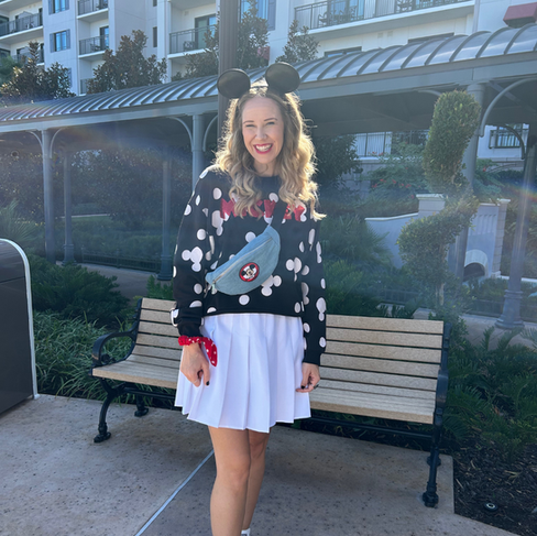 Ashlie from Once Upon a Wardrobe standing in front of the Disney's Riviera wearing a mickey mouse sweater, white skirt, black ears, and a mickey fanny pack