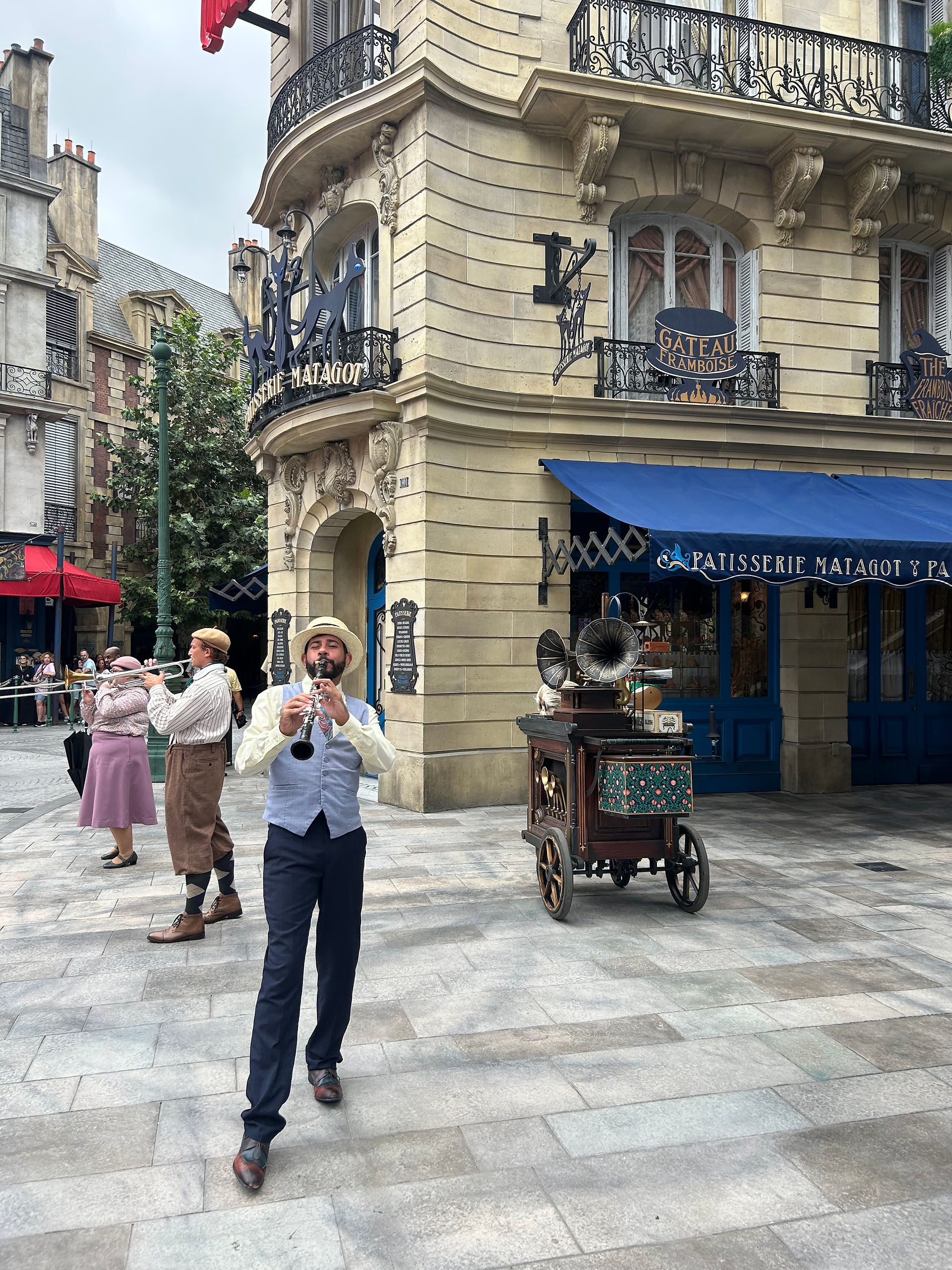 Street performers entertaining guests in the Paris-themed Wizarding World at Epic Universe, with musicians performing in period costumes near French wizarding storefronts.