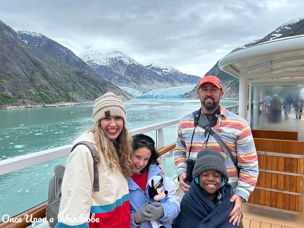 A smiling family poses on the deck of a Disney Alaska cruise ship with a glacier and snow-capped mountains in the background. The group is bundled in winter layers, including a beanie, gloves, and Mickey-themed gear, with binoculars and a Captain Mickey plush in hand. The teal water and icy landscape highlight the breathtaking Alaskan scenery.