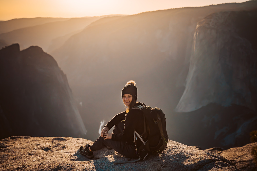 A elopement photographer wearing a hiking backpack sitting on a rock overlooking the sunset on the top of Yosemite Valley