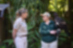 Two zookeepers in uniforms converse in a lush, green setting. One holds a large bird of prey. Text on uniforms: "Australia Zoo."