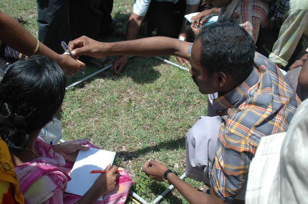 Group of people sit on grass outdoors, passing a pen and taking notes in notebooks during a discussion.