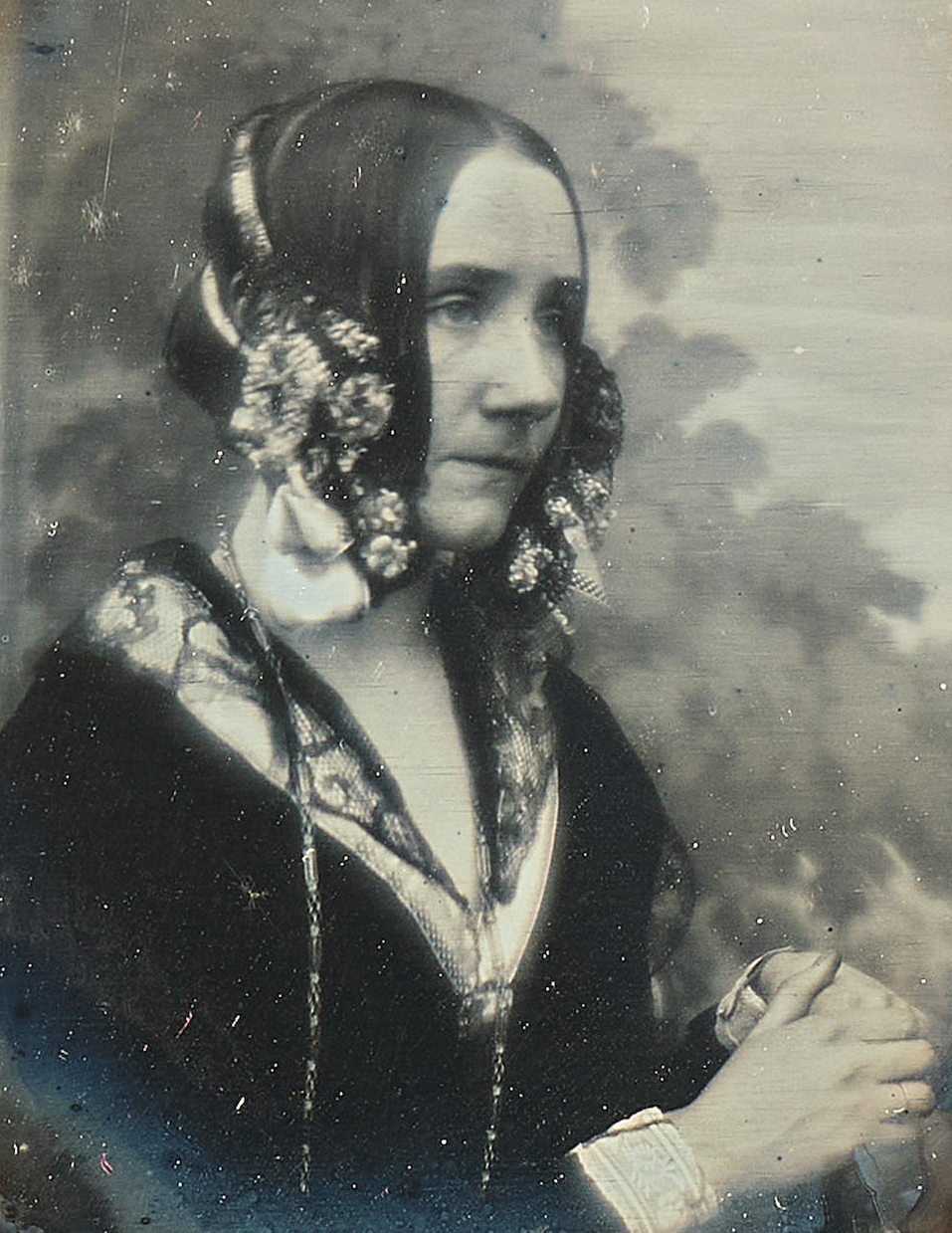 Woman in vintage attire with floral headpieces gazes to the side, holding a small object. Sepia tones evoke a calm, historical mood.