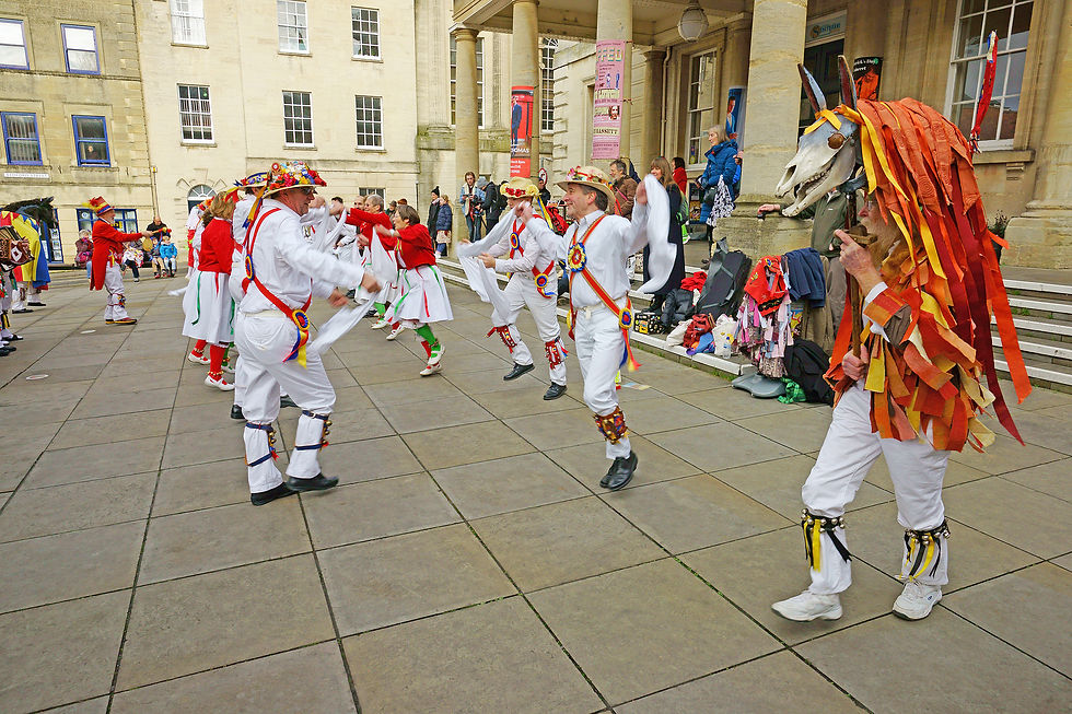 Morris dancers in colorful outfits perform outdoors. One dancer wears a horse costume. Spectators watch from building steps. Festive mood.
