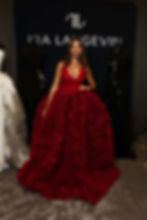 Sofia Valleri in a red, floral gown stands confidently between black mannequins. Background features "IRA LANGEVIN" text on a dark backdrop. Luxurious setting.