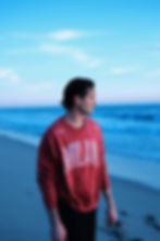 Noah Fearnley in red "MILAN" sweater stands on a beach, looking at the ocean under a clear blue sky. Peaceful, contemplative mood.