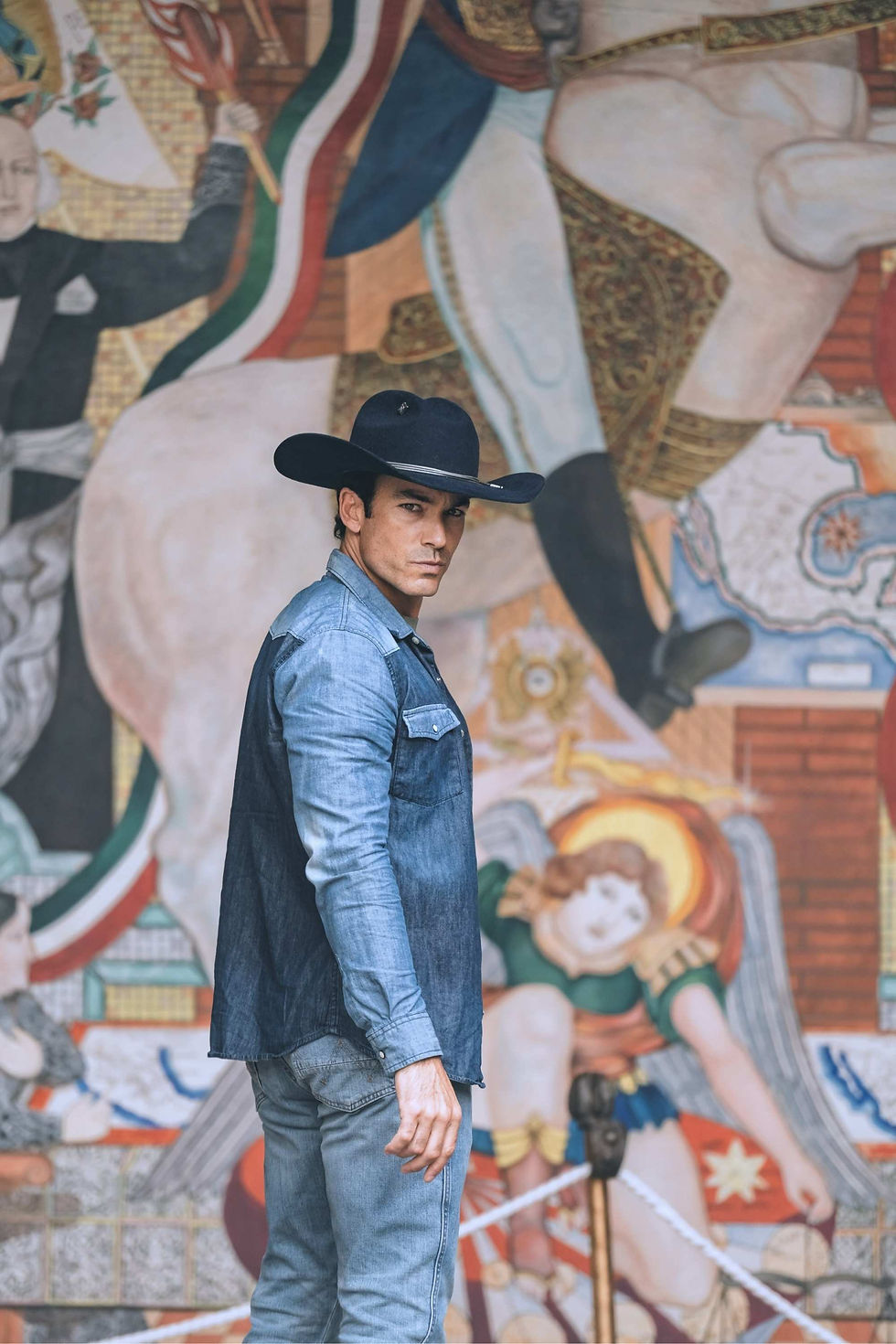 Aarón Díaz in a black cowboy hat and denim outfit stands in profile before a colorful mural, looking at the camera with a serious expression.