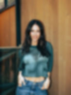 Smiling Jessica Lowndes with long dark hair poses against a wood-paneled wall, wearing a teal mesh top and blue jeans.