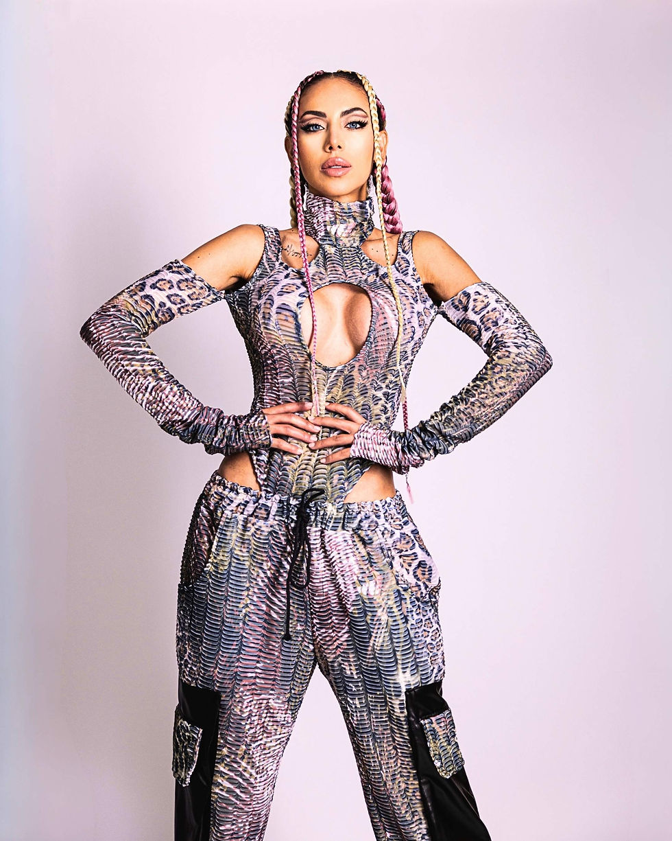 Esther Anaya in a patterned, cutout outfit with braids stands confidently, hands on hips. Pale background enhances vibrant attire.