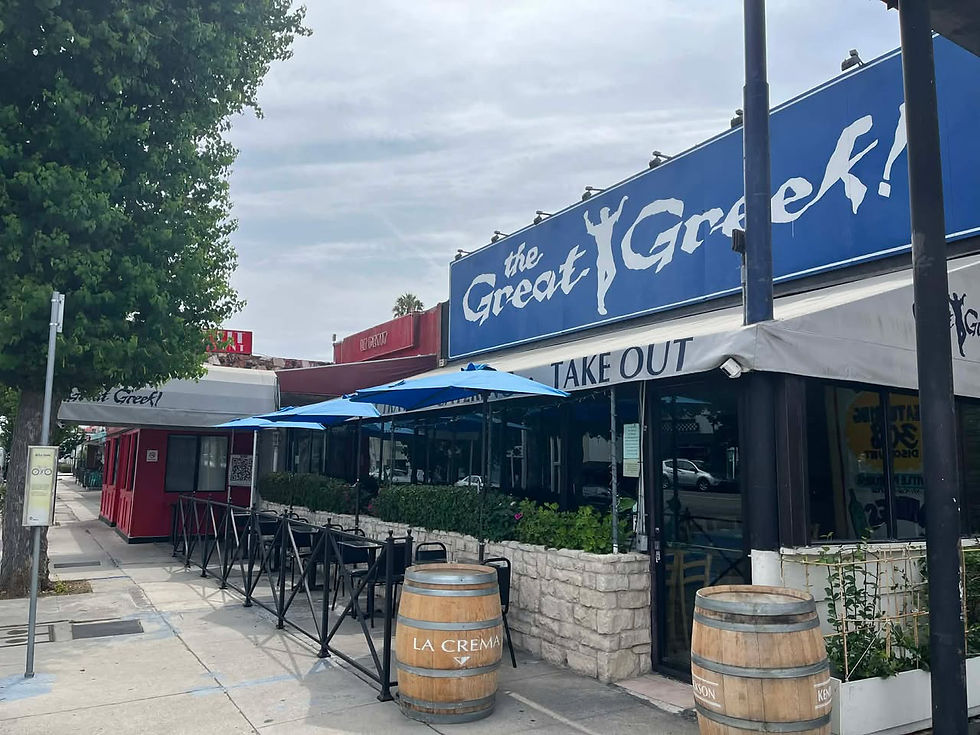 Street view of "The Great Greek" restaurant with blue umbrellas and barrels outside. Sign reads "TAKE OUT." Greenery accents exterior.