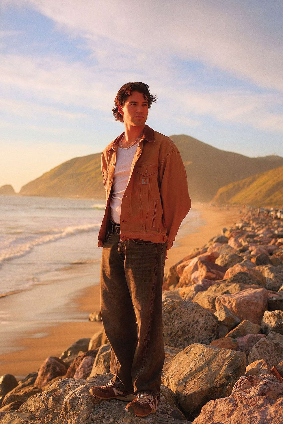 Noah Fearnley stands on rocky shore at sunset, wearing a brown jacket and jeans, with mountains and ocean in the background. Calm atmosphere.