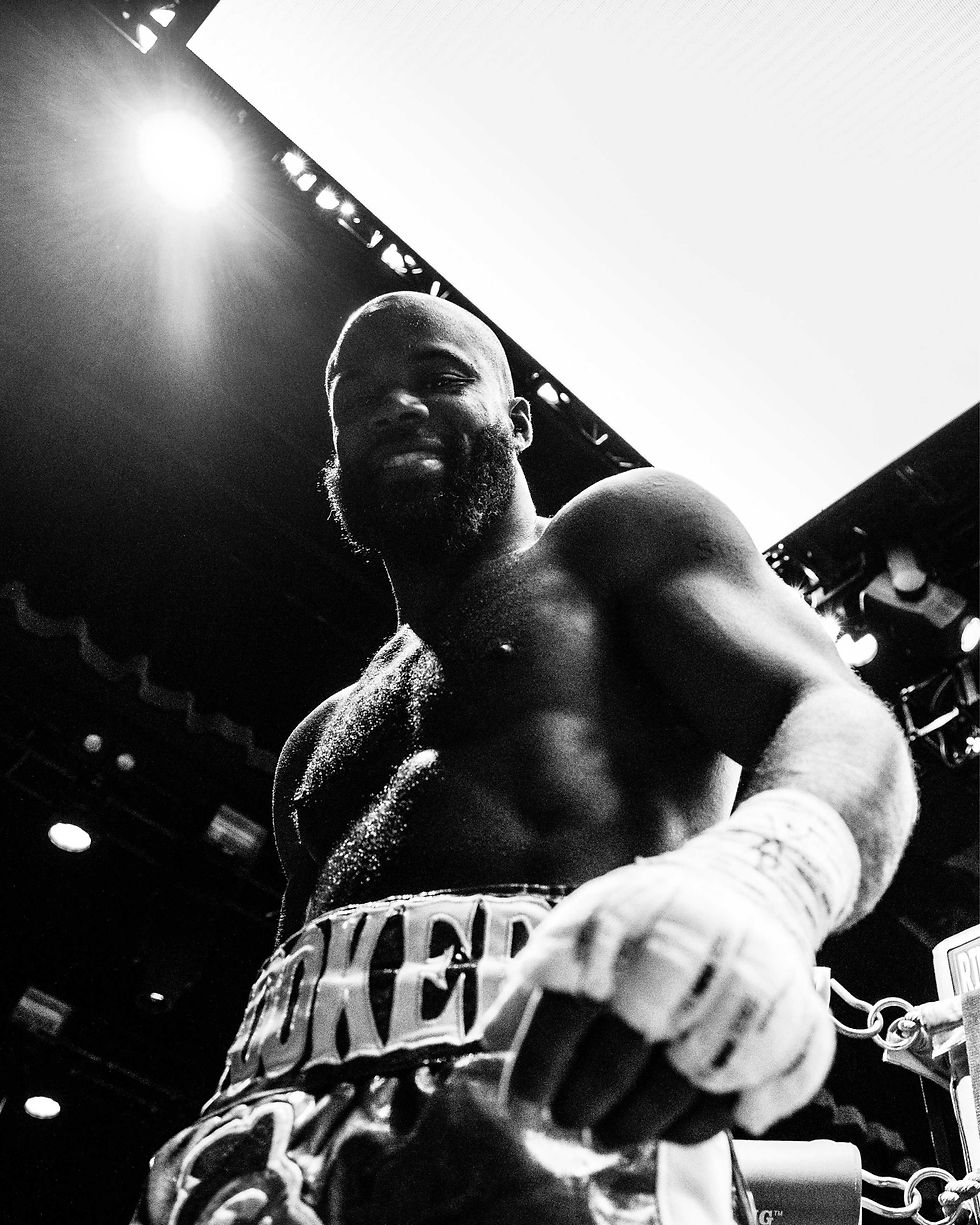 Metta World Peace with boxer gloves stands confidently in a ring under bright lights. His shorts display text. The black-and-white image exudes determination.
