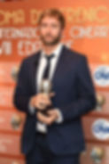 Paki Meduri in a navy suit holds a trophy at a Roma cinema awards backdrop, before orange banners with event logos.