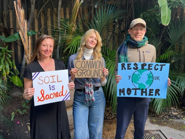 Rebecca Tickell, Laura Dern, and Josh Tickell in ‘Common Ground.’ Photo Courtesy of Big Picture Ranch hold signs reading "Soil is Sacred," "Common Ground," and "Respect Your Mother." They stand in a lush garden, smiling.