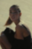 Sofia Valleri with a ponytail crouches outdoors on a sunlit tiled surface, wearing a black dress. She's looking at the camera with a calm expression.