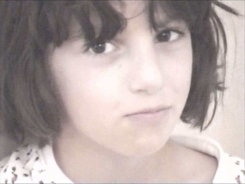Young Jihan with dark hair and bangs looks at the camera with a neutral expression. She wears a white shirt with black patterns. Neutral background.