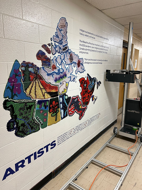 Direct-to-wall printed mural on an interior concrete block school wall depicting a detailed map of Canada filled with colorful indigenous beadwork designs, alongside explanatory text about the BeadYourProvince movement and listed artist names, with a vertical wall printing machine visible on the right.