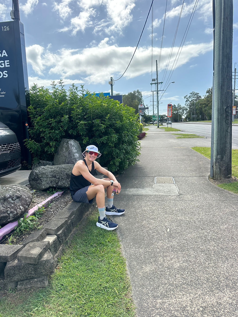 Person sitzt auf einem Bordstein neben Sträuchern, trägt Hut und Sonnenbrille. Straße und bewölkter Himmel im Hintergrund. Entspannt.