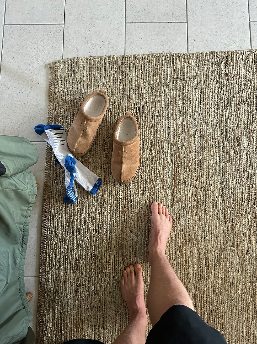 Blick von oben auf nackte Füße auf einem geflochtenen Teppich, daneben Pantoffeln und blaue Socken auf hellem Fliesenboden.