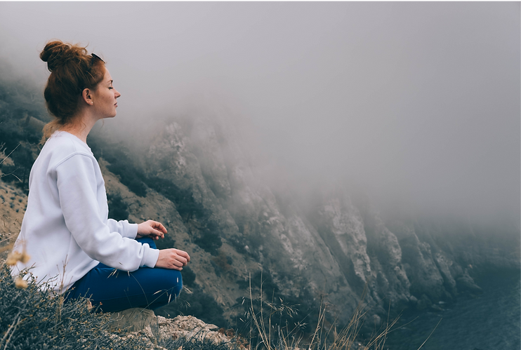 Kvinna i vit tröja sitter i meditativ ställning på en klippavsats i dimhöljda berg, med slutna ögon och avslappnat ansiktsuttryck
