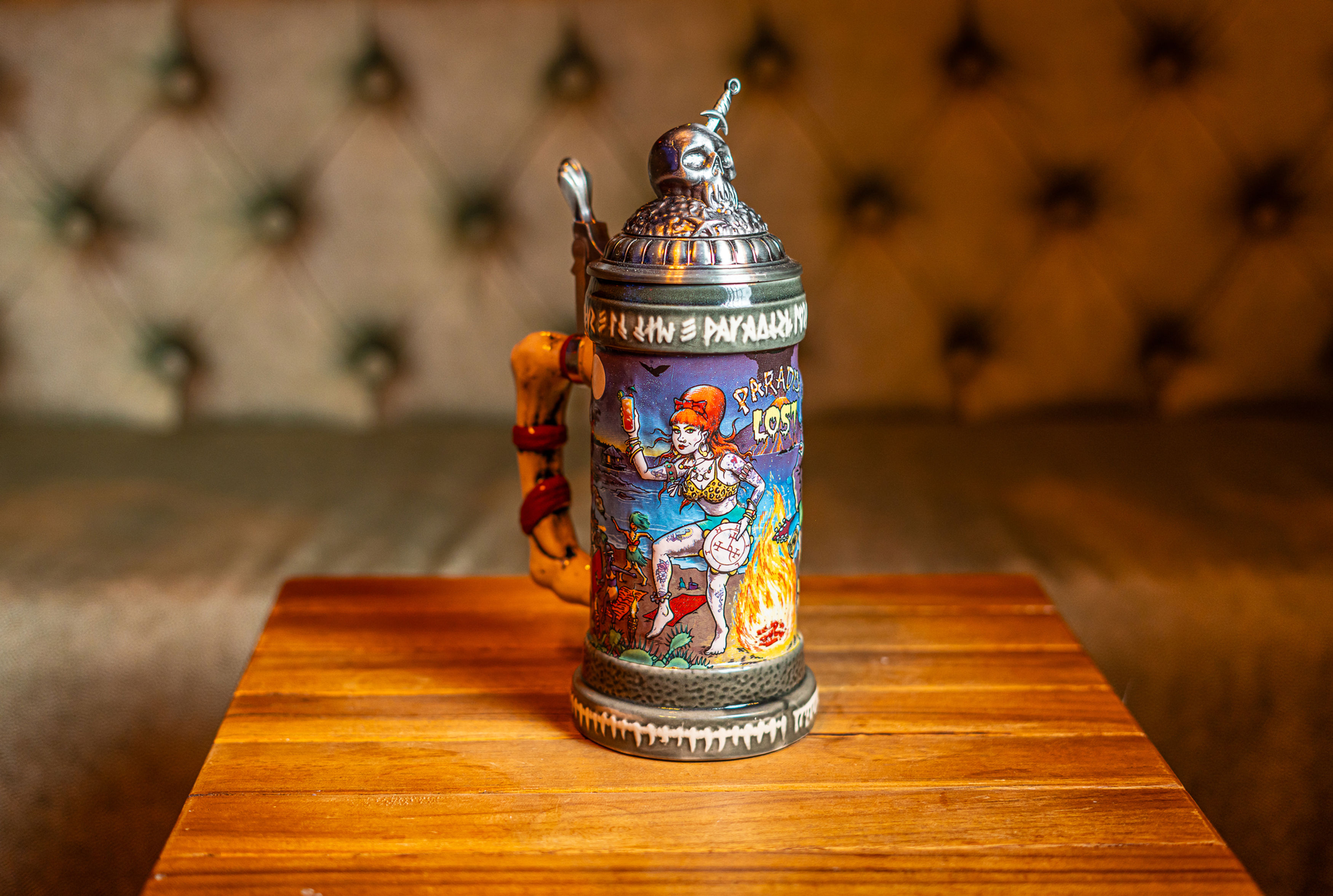 A stein sits upon a wooden table. The stein depicts Lilith and Beelzebub partying around a bon fire among other demons. This image depics the left side of the stein.