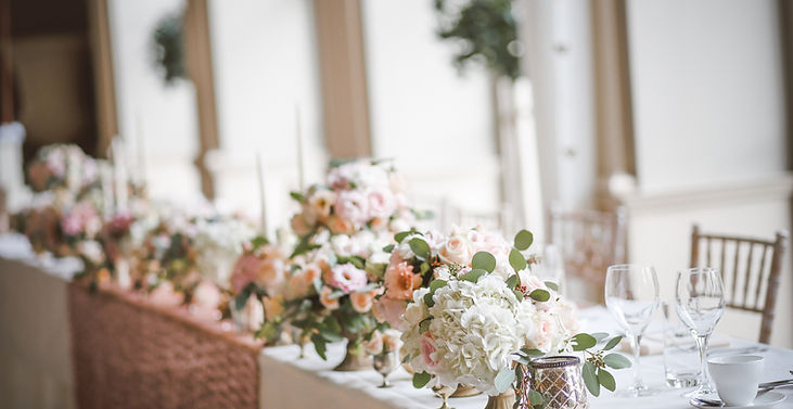 A long table set for a special occasion, with white tablecloths, white chairs, and three centerpieces of white flowers and candles.