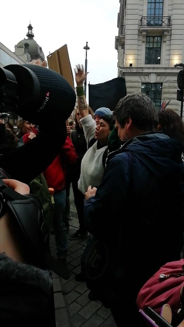 A protestor in a white jumper raises a hand as they are about to shout a chant. They are in the midst of a tight crowd of protestors. A professional camera microphone pokes out from the left of the image.