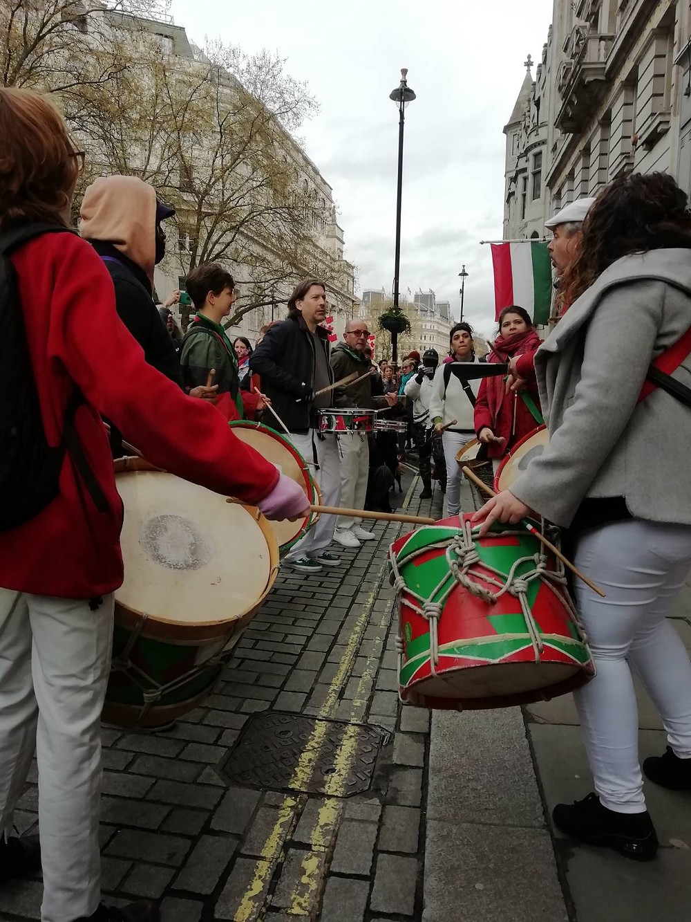 Drummers from Baque de Axé line the street. The italian flag from a distant embassy is seen in the background. Their drums are green and red patterned, supported by bands of rope, and are held to the upper thigh. They are roughly the size of a large waste paper basket.