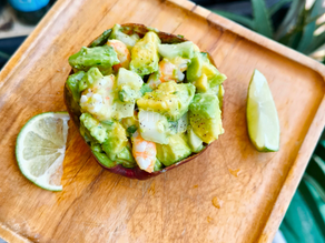 Assiette en bois carrée avec un demi avocat facis avec des mroceaux de concmbre, avocat et crevettes et deux morceaux de citorn vert